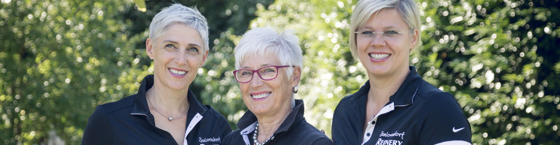 Drei lächelnde Führungskräfte der Reinery Pflege GmbH in Dienstkleidung vor grünem Hintergrund.