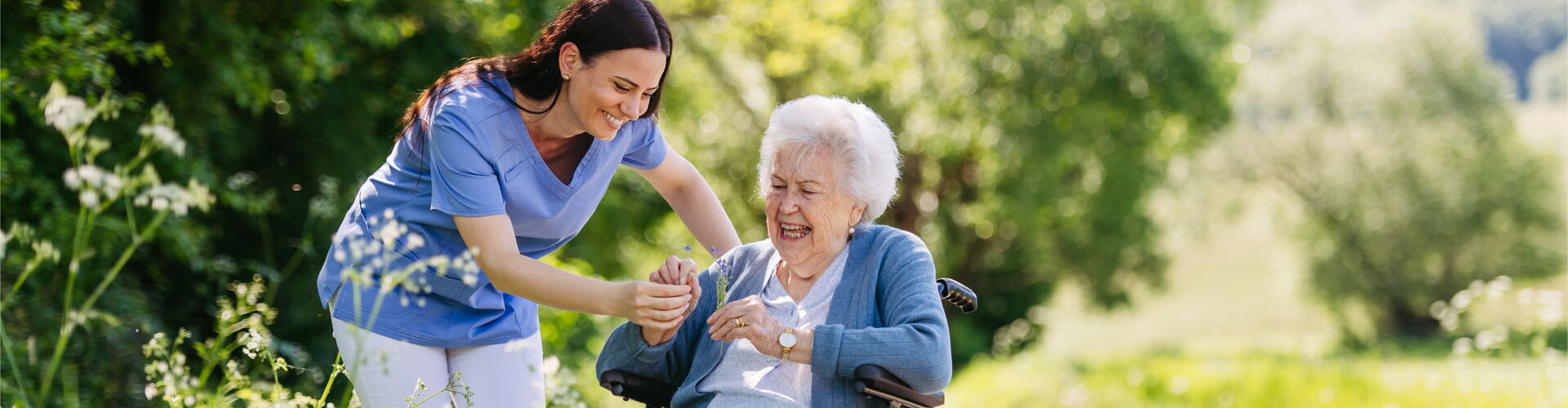 Eine Pflegekraft begleitet eine Seniorin im Rollstuhl bei einem sonnigen Ausflug in die Natur – Gemeinsames Erleben und liebevolle Betreuung.